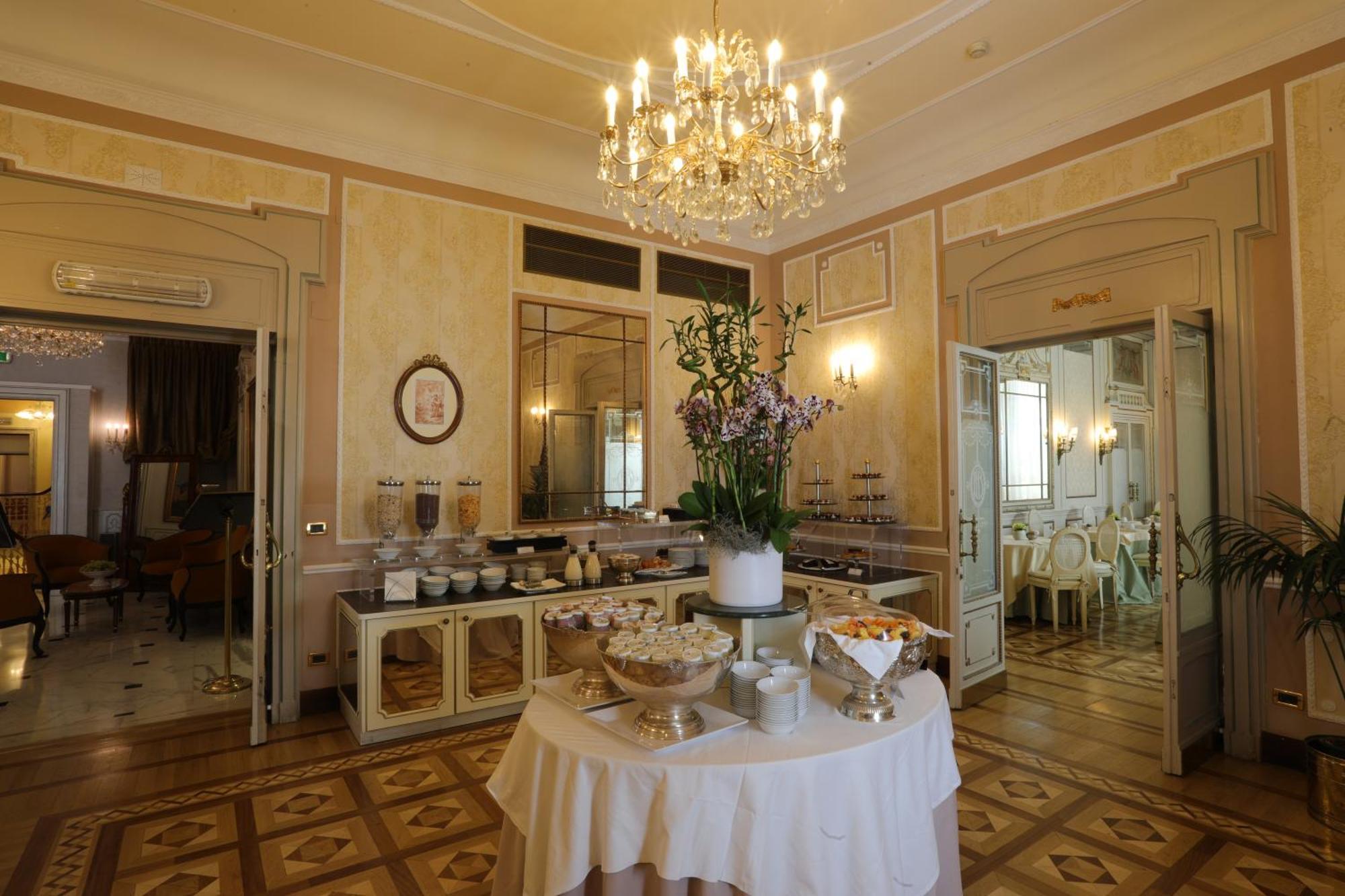 Hotel Bristol Palace Genoa Bagian luar foto The photo shows an elegant interior of a dining area or event space. There is a beautifully decorated chandelier hanging from the ceiling, illuminating the room. In the center, there's a table draped with a white tablecloth, featuring a display of va